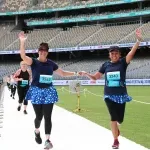 Two women with their arms raised in celebration run towards the camera, holding hands, with a stadium in the background.