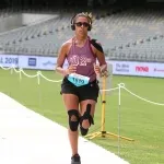 A female runner wearing headphones and knee braces runs on a paved path with stadium seating in the background.