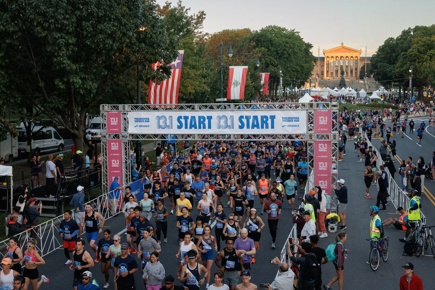 Runners surge forward under the '13.1 START' banner at the Philadelphia Distance Run, with the Philadelphia Museum of Art visible in the background.