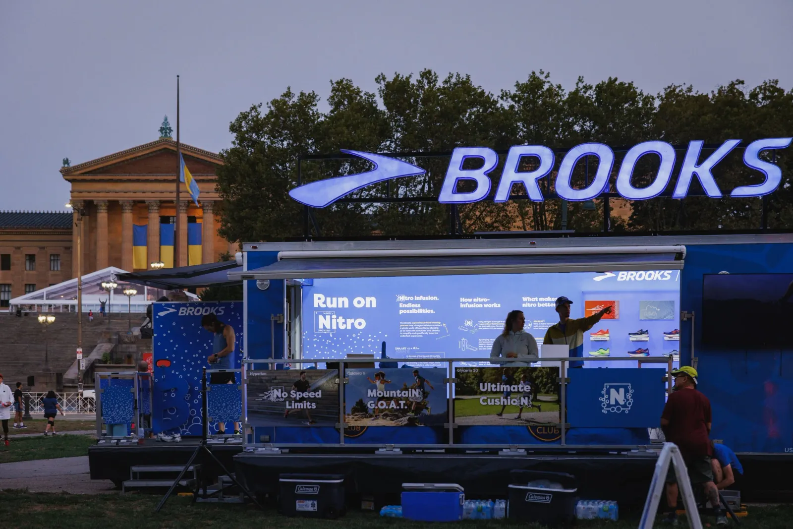 People interacting with a Brooks running booth at a race event, with the Philadelphia Museum of Art in the background.