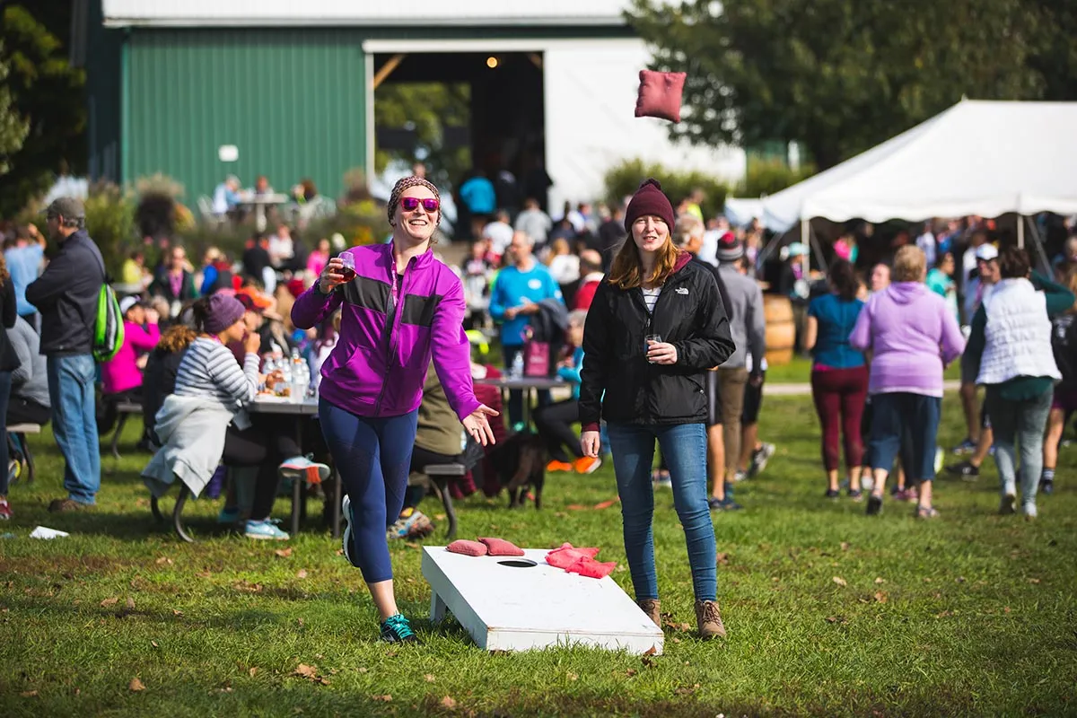 Two women smiling and playing a beanbag toss game at an outdoor festival with a crowd in the background.