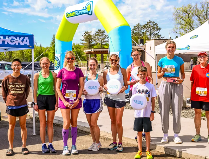 A group of runners, including men, women, and a child, pose for a photo under a colorful inflatable archway at a race event. Some are holding frisbees, and bib numbers are visible on some participants.