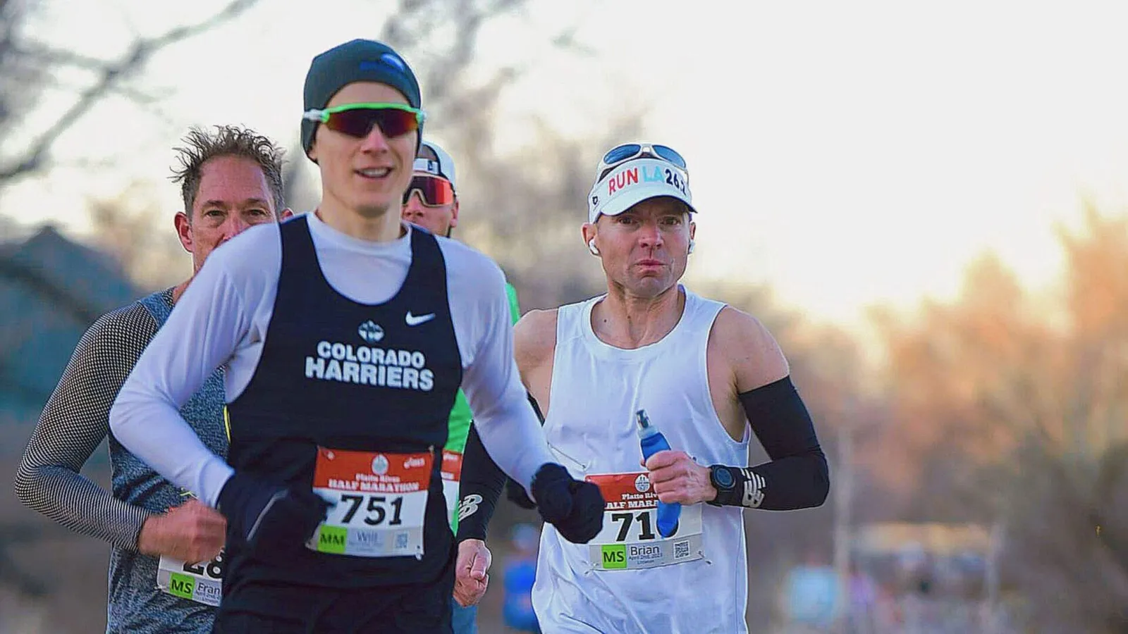 Runners competing in the Platte River Half Marathon, with one runner in the foreground wearing a 'Colorado Harriers' singlet.