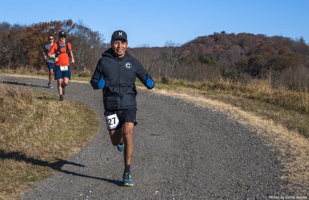 A runner wearing bib number 27 smiles and gives a thumbs up while running on a gravel path, with other runners behind him and a wooded hillside in the background.