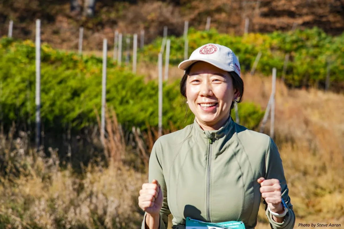 A smiling woman wearing a race bib and a cap, with her fists clenched in excitement, outdoors on a sunny day.