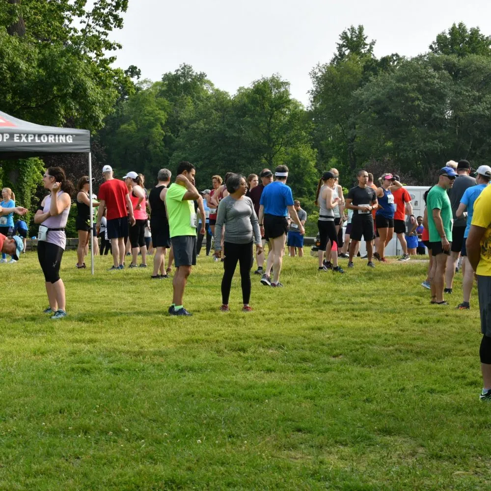 A group of runners standing on a grassy field before a race, with trees and a tent in the background.