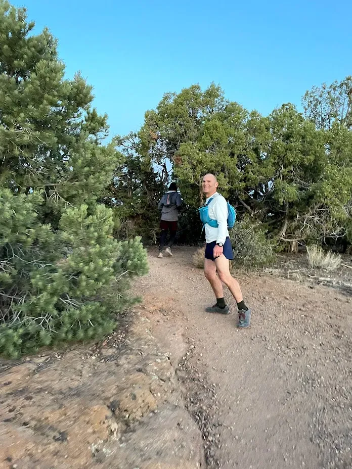 A runner smiles at the camera while running on a desert trail, with another runner in the background.