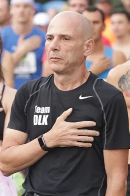A bald man in a black Nike shirt with 'team DLK' on it places his hand over his heart while standing in a crowd of runners at the start line.