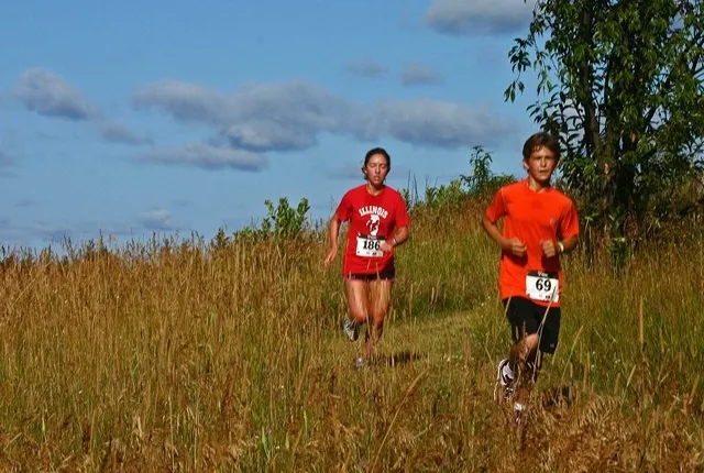 Two young runners, a girl in a red shirt and a boy in an orange shirt, running through tall grass on a sunny day.