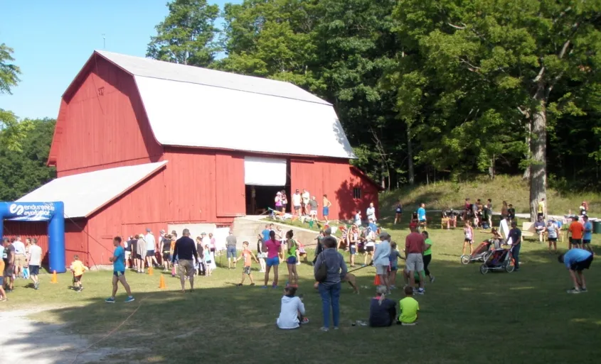 Runners and spectators gathered near a red barn with an inflatable arch at the start or finish line of a race.