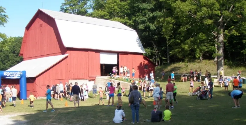 A group of runners and spectators gather near a red barn with an inflatable start/finish arch at the Port Oneida Run.