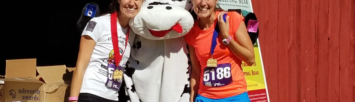 Two smiling female runners wearing medals pose with a cow mascot at a race finish line.