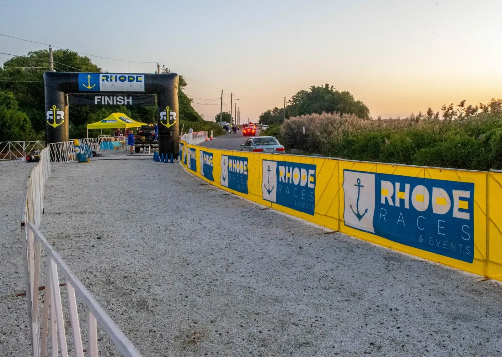 Inflatable archway with 'FINISH' text and 'Rhode Races & Events' branding, set up at the start/finish line of a race, with yellow sponsor banners lining the course.
