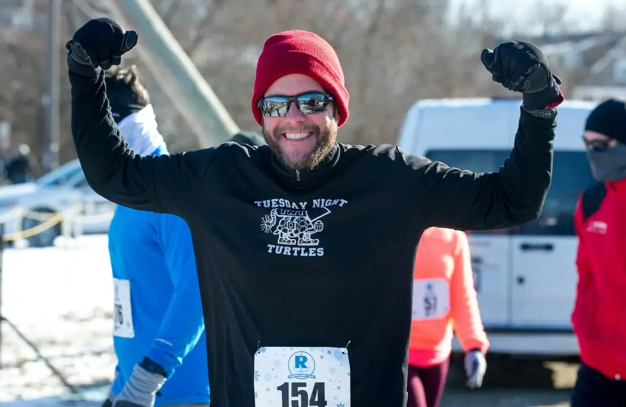 A smiling runner in a red beanie and sunglasses flexes his arms in celebration at a race.