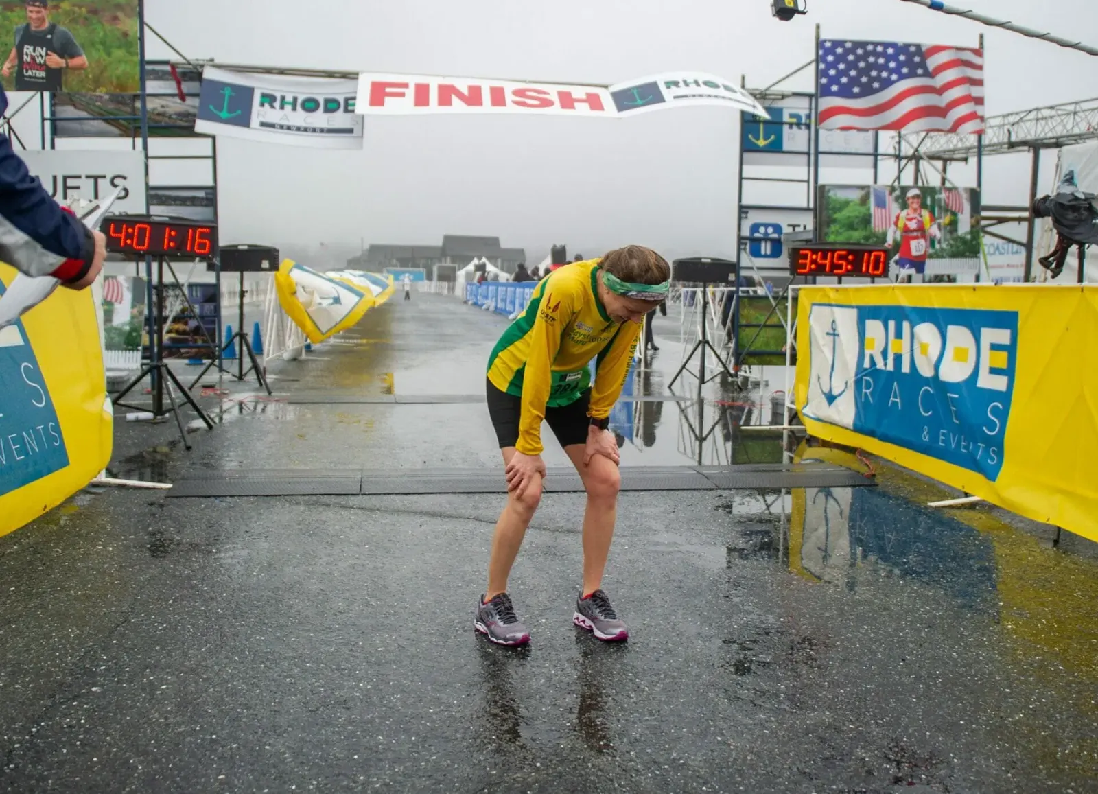 A runner bends over, catching their breath at the finish line of a race on a wet, overcast day.