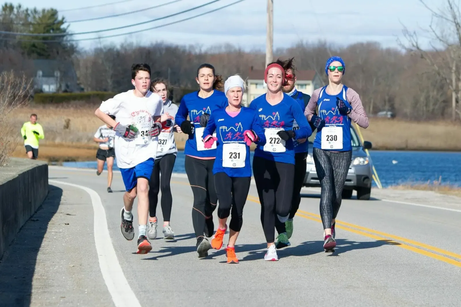 A group of runners, including a young male and several women, run along a road with a body of water in the background.