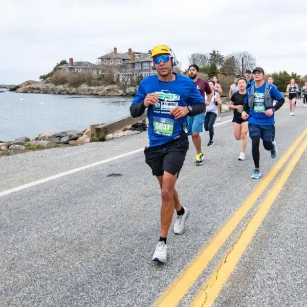 A runner in a blue shirt and yellow hat runs along a road next to the water, with other runners behind him.