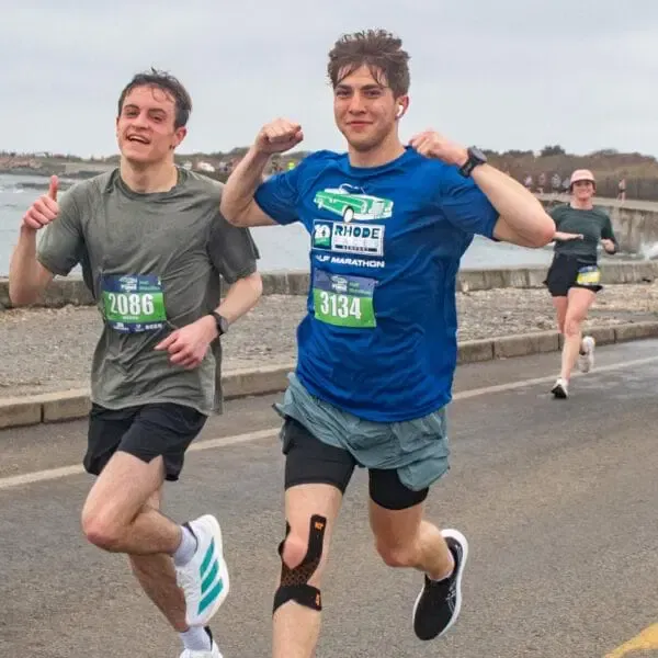 Two male runners, one giving a thumbs up and the other flexing his biceps, run along a road with a waterfront in the background.