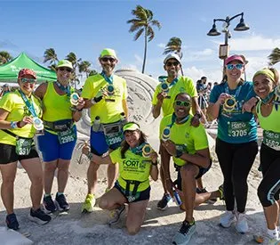 A group of eight runners, some wearing neon yellow shirts and others in blue shorts, pose for a photo with their race medals around their necks. They are standing in front of a sand sculpture, with palm trees and a bright blue sky in the background.