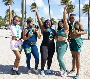 A group of five runners, all wearing medals, pose for a photo on a beach with palm trees in the background.