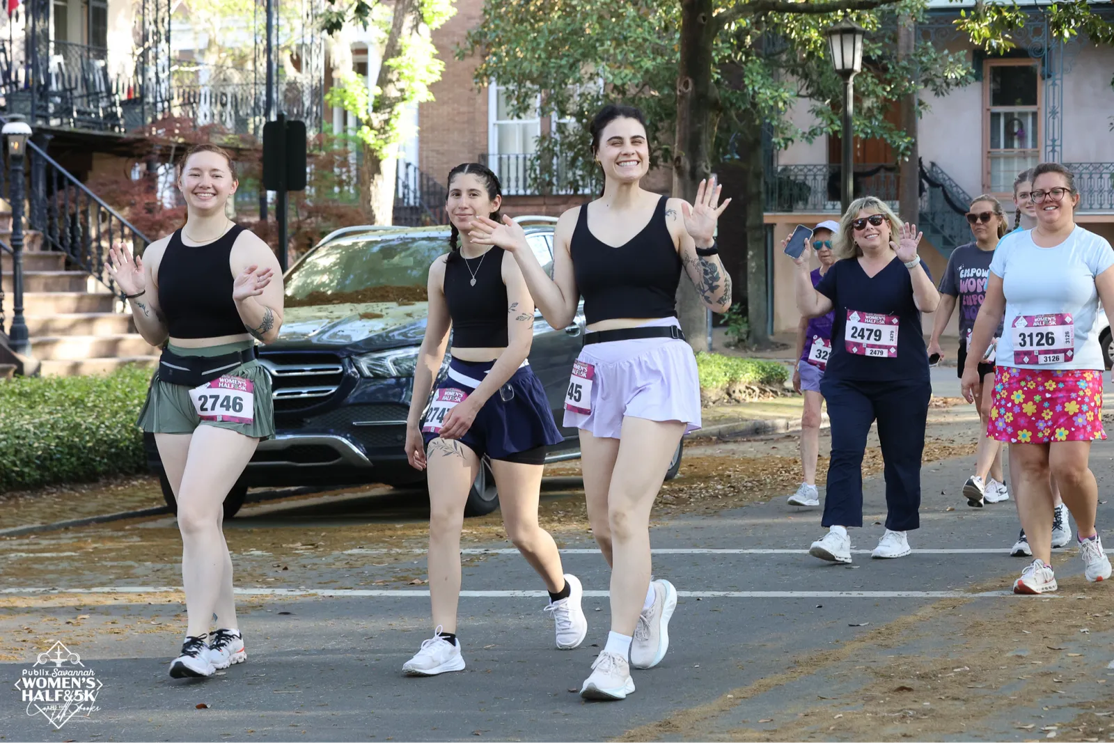 A group of women smiling and waving as they run in a race, with historic buildings in the background.
