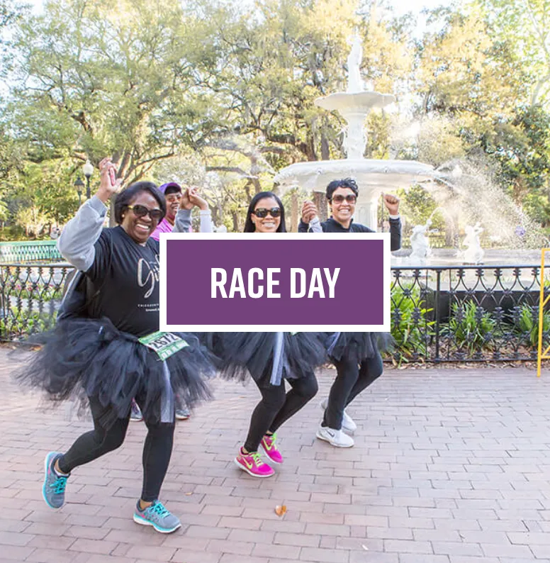 Four women in black tutus and running attire smile and cheer while running in front of a fountain, with a purple banner that reads 'RACE DAY'.