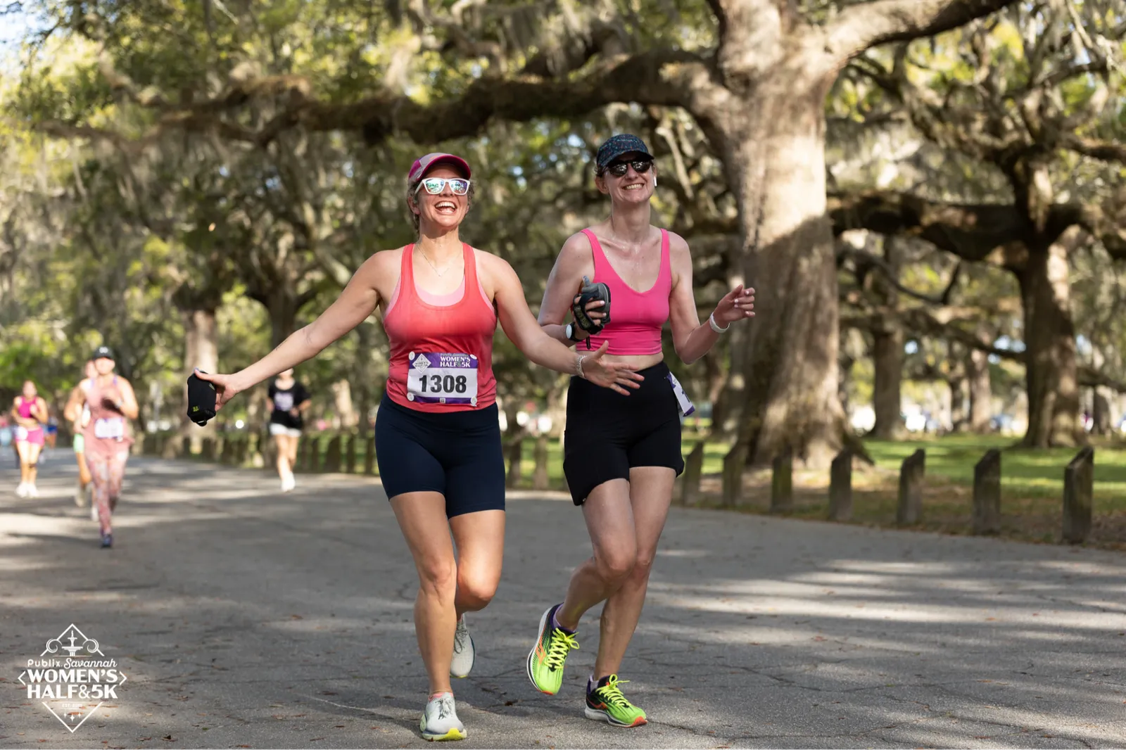 Two women running and smiling during the Publix Savannah Women's Half Marathon & 5K, with large oak trees and Spanish moss lining the course.