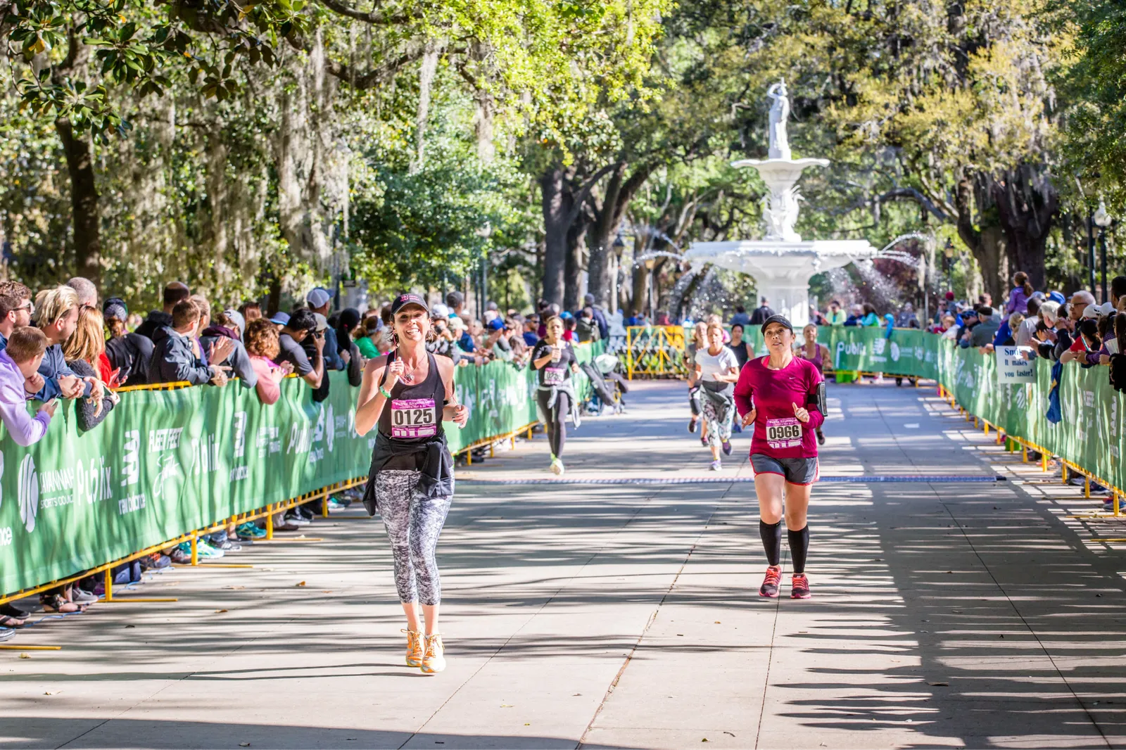 Runners smiling as they race down a tree-lined street with spectators cheering from behind a green barrier, a fountain visible in the background.