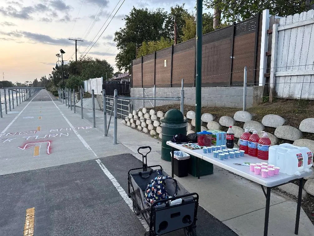 A paved waterfront path with yellow and white lane markings, a refreshment table with drinks and cups, and a trash can.