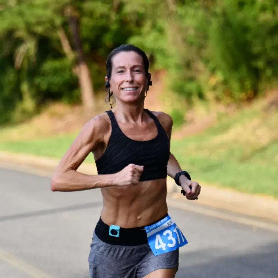 A female runner with a bib number 431 smiles as she runs on a paved road with trees in the background.