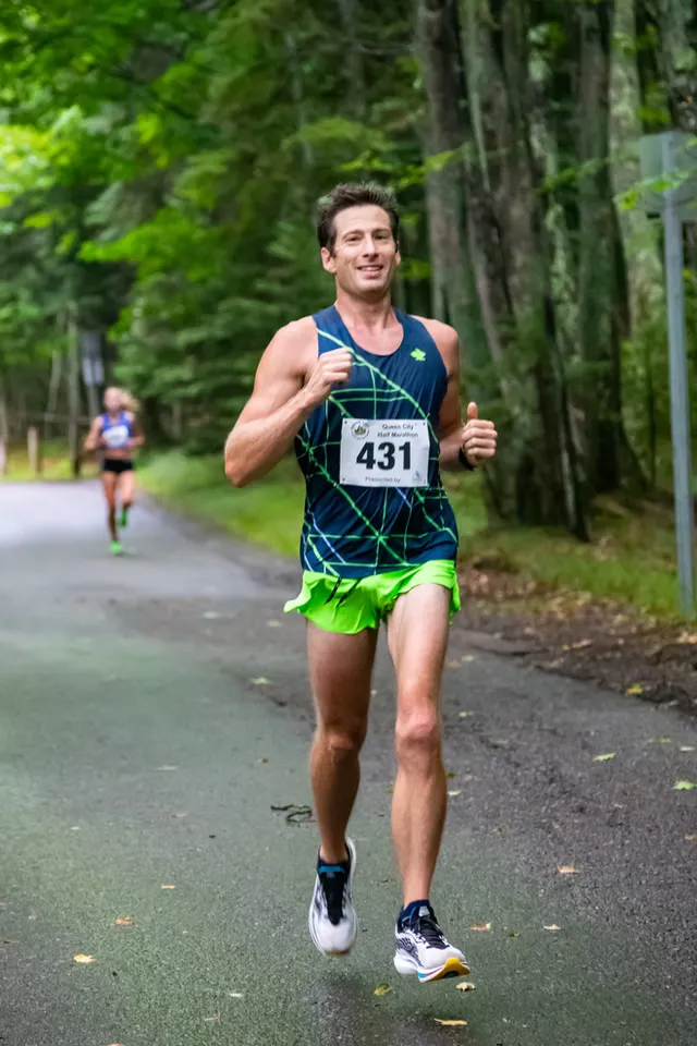 A male runner with bib number 431 smiles as he runs on a paved road through a forest.