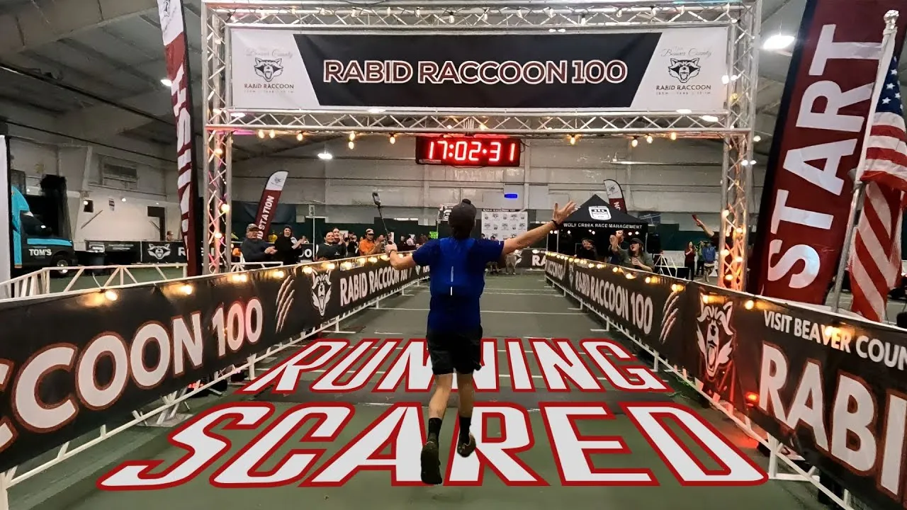 A runner crosses the finish line with arms raised in celebration at the Rabid Raccoon 100 race, with a crowd cheering and race banners visible.