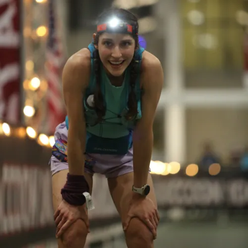 A female runner with a headlamp, braids, and a determined expression, bent over with hands on her knees, appearing to have just finished a race.