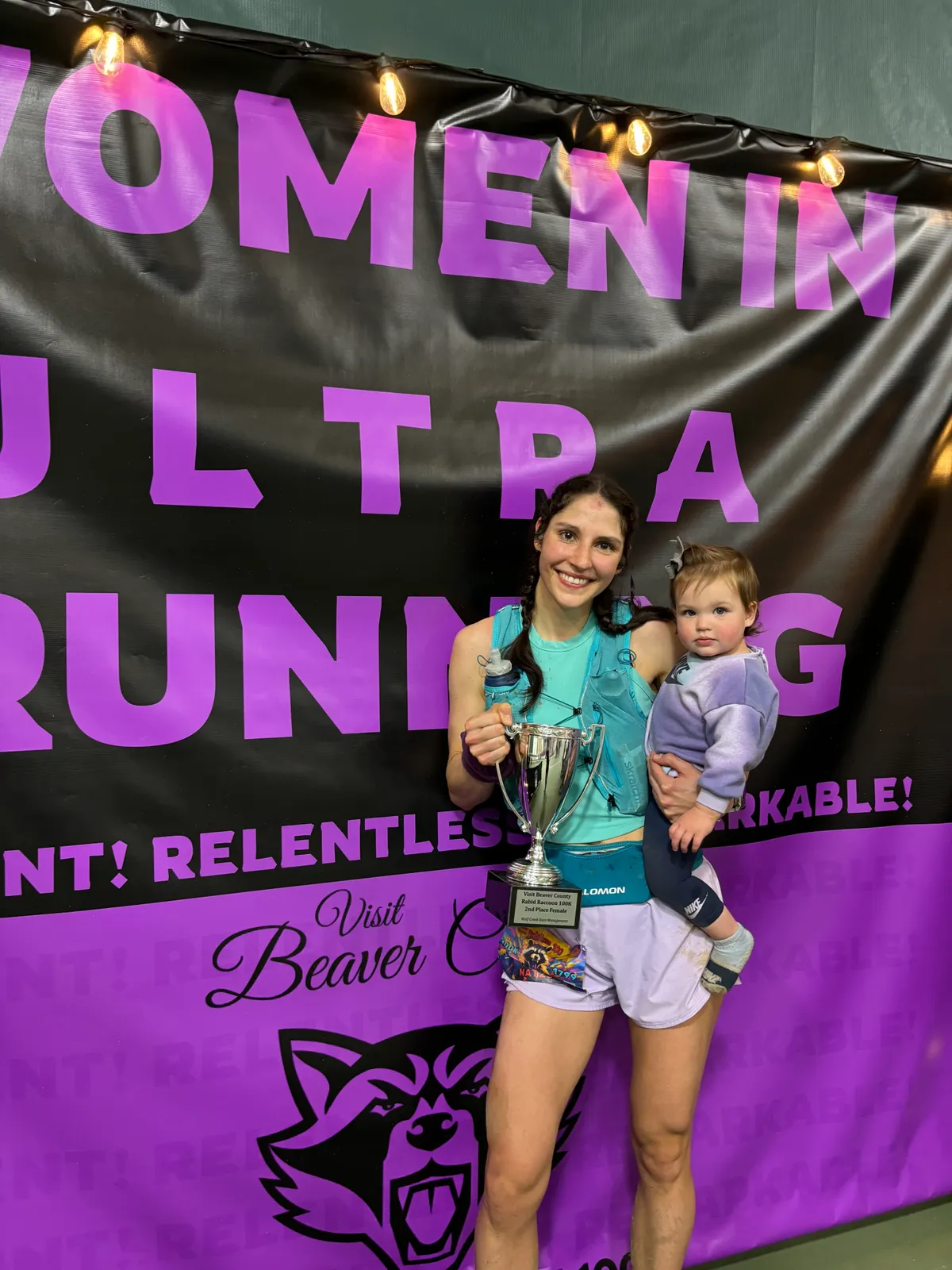 A female runner smiles while holding a trophy and a child in front of a banner that says 'WOMEN IN ULTRA RUNNING'.