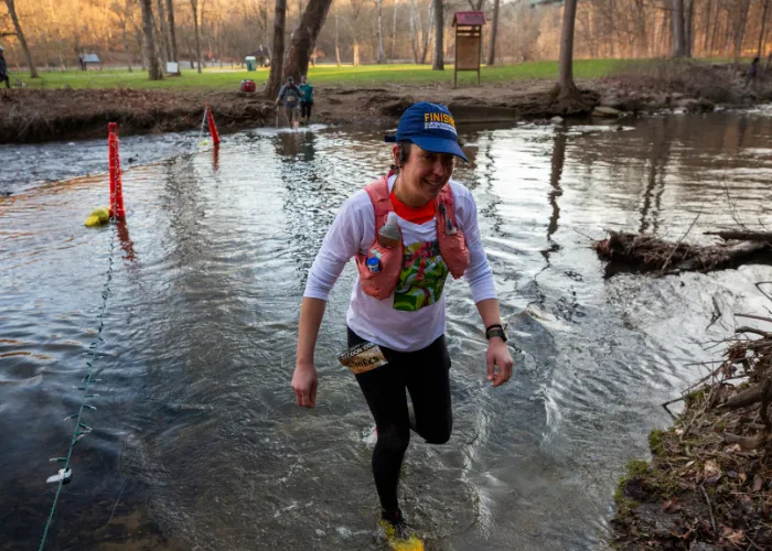A runner smiles as they wade through a shallow stream during a race, wearing a blue 'Finish' hat and a hydration vest.