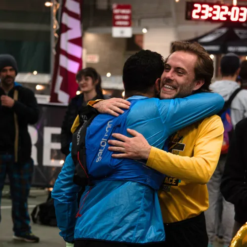 Two runners embrace and celebrate at a race, with a digital clock showing 23:32:03 in the background.