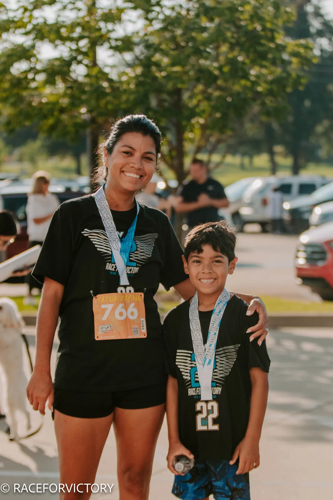 A smiling woman and a young boy, both wearing race t-shirts and medals, pose for a photo after a race.