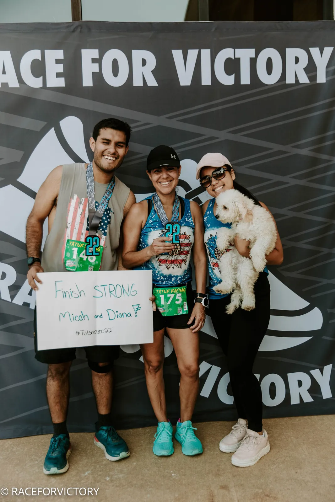 Three smiling people, one holding a dog, pose in front of a 'Race For Victory' banner, all wearing race medals and bibs.