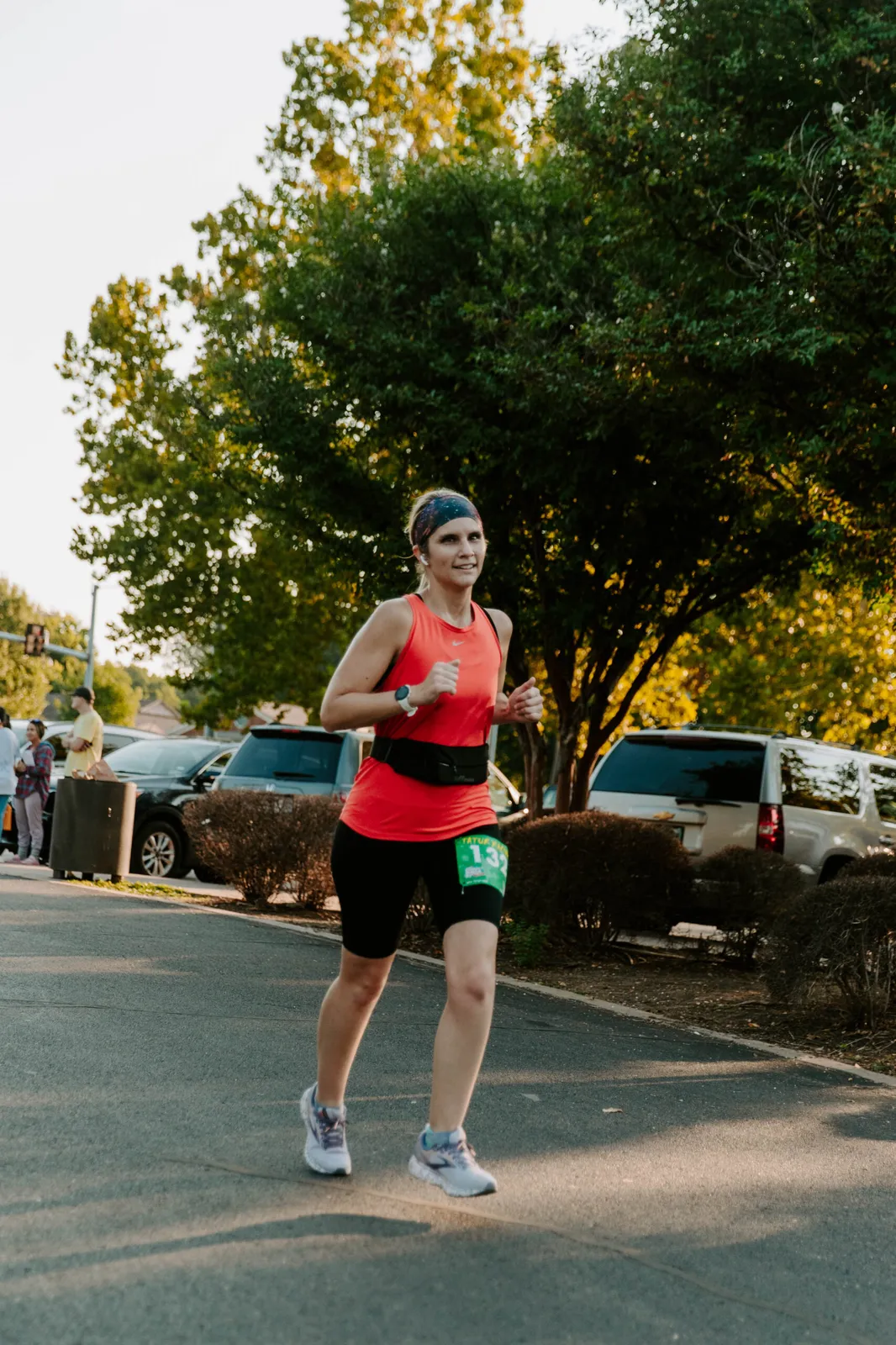 A female runner in an orange tank top and black shorts runs on a paved road, with trees and cars in the background.