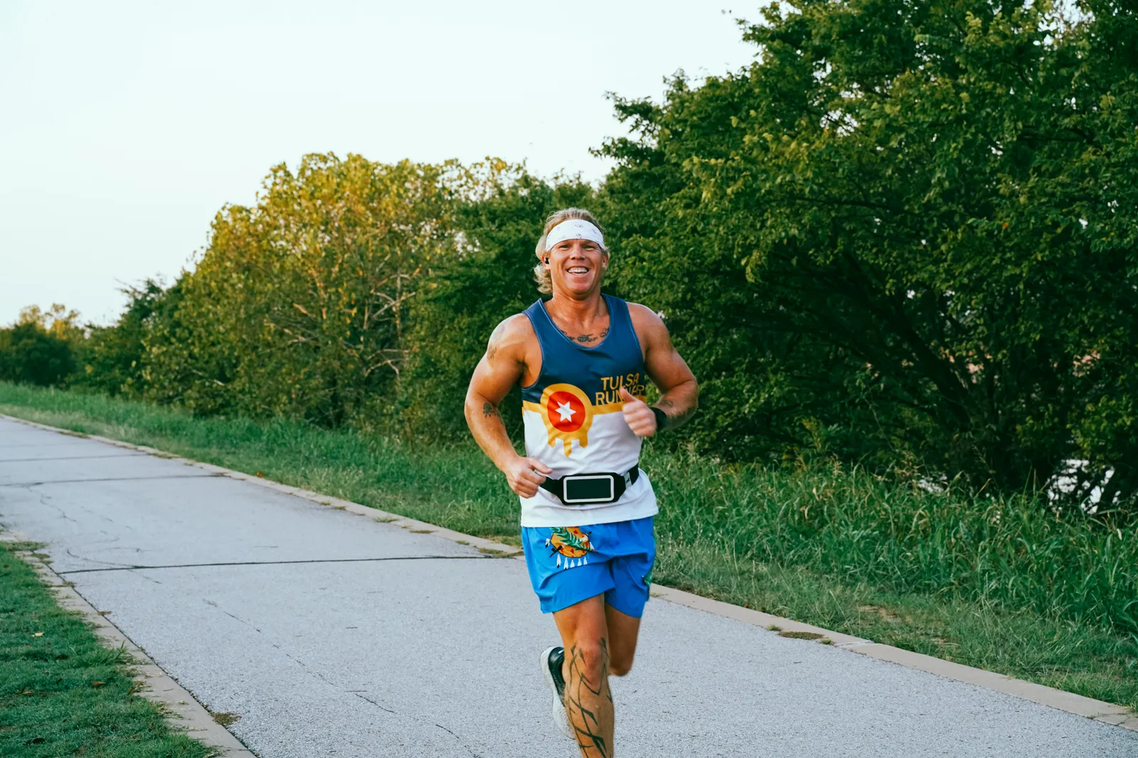 A male runner wearing a Tulsa Runner singlet and blue shorts smiles as he runs on a paved path lined with trees and grass.