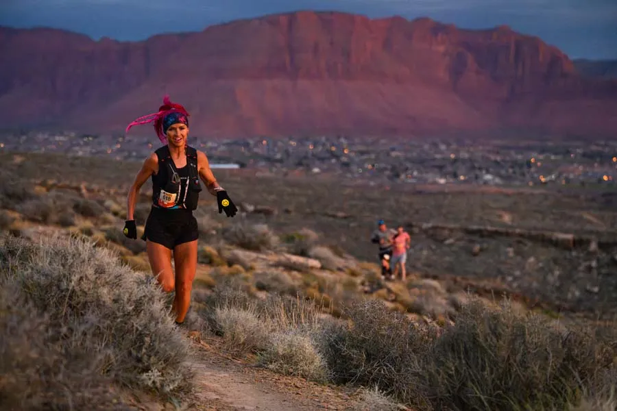 A female runner with pink hair runs down a dusty trail with a scenic mountain and city lights in the background at dusk.