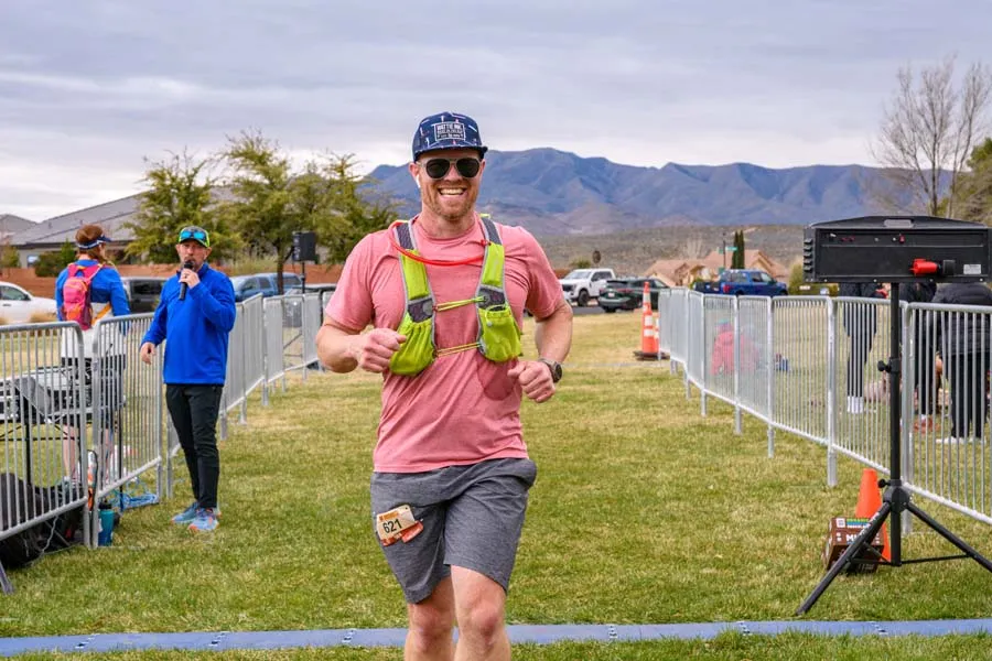 A smiling male runner wearing a pink shirt, grey shorts, and a hydration vest runs towards the camera with mountains in the background.