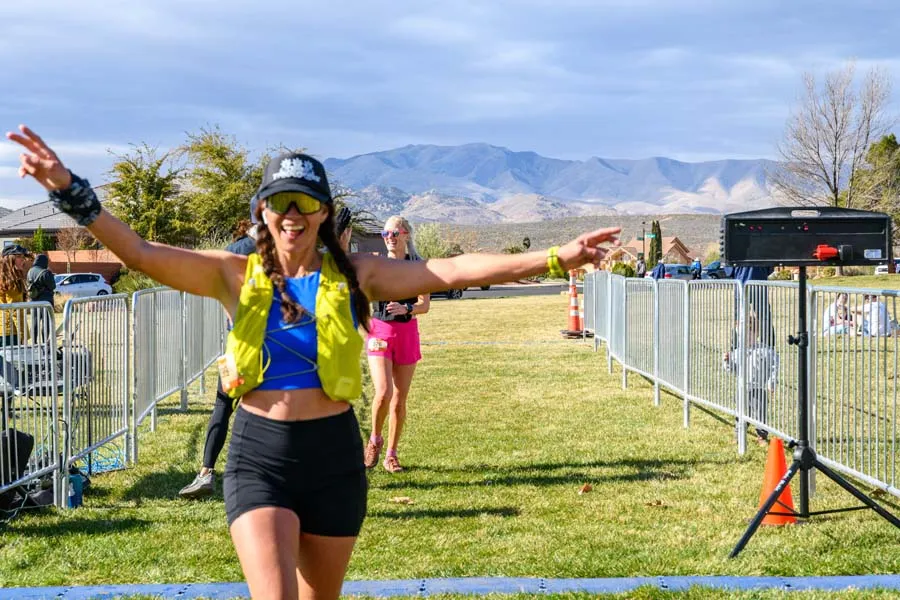 A female runner with arms outstretched and a peace sign, celebrating as she crosses the finish line of a race, with mountains in the background.