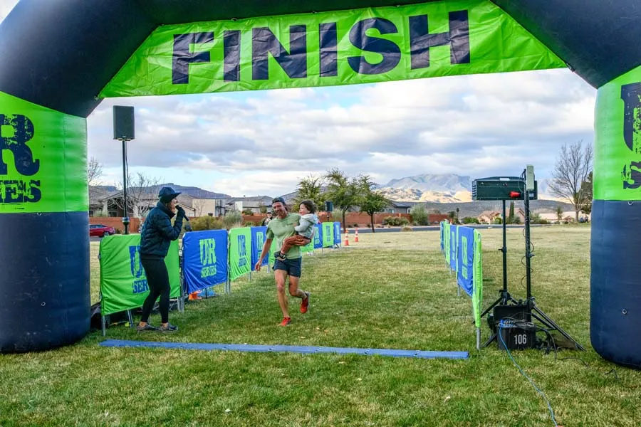 A runner carrying a child crosses the finish line under a green and black inflatable arch.