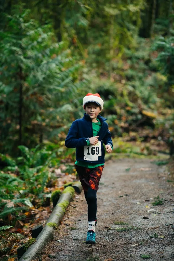 A young runner wearing a Santa hat and bib number 169 runs on a forest trail.