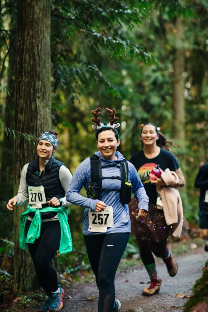 Three women running on a wooded trail, two wearing reindeer antlers.