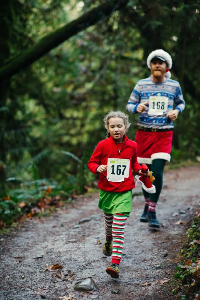 A young girl in a red top and green skirt, wearing striped socks and a race bib number 167, runs on a forest trail, followed by an adult in a Santa hat and festive sweater with bib number 168.