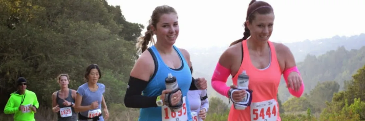Two women running a trail race, smiling at the camera, with other runners and a scenic, hilly background.