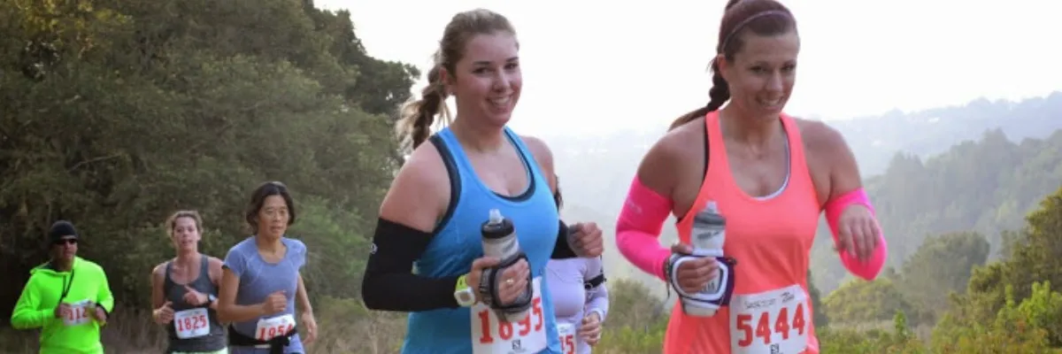 Runners smiling as they participate in a trail race, with a scenic, hilly landscape in the background.