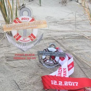 Two Rehoboth Beach Running Company marathon medals displayed on sand, one larger with an anchor and life preserver design, the other smaller with a life preserver design. The larger medal indicates '2017 10th Anniversary Marathon' and 'First Place Female 30-34'. The smaller medal also shows '10th Anniversary Marathon'. A red ribbon with '12.2.2017' is also visible.